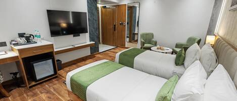 A hotel room with two beds, a desk with a computer, a flat-screen TV, a mirror, and a green armchair.