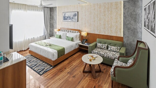 Habitación de hotel con cama, sofá verde, mesa de centro redonda, escritorio con lámpara y televisor.