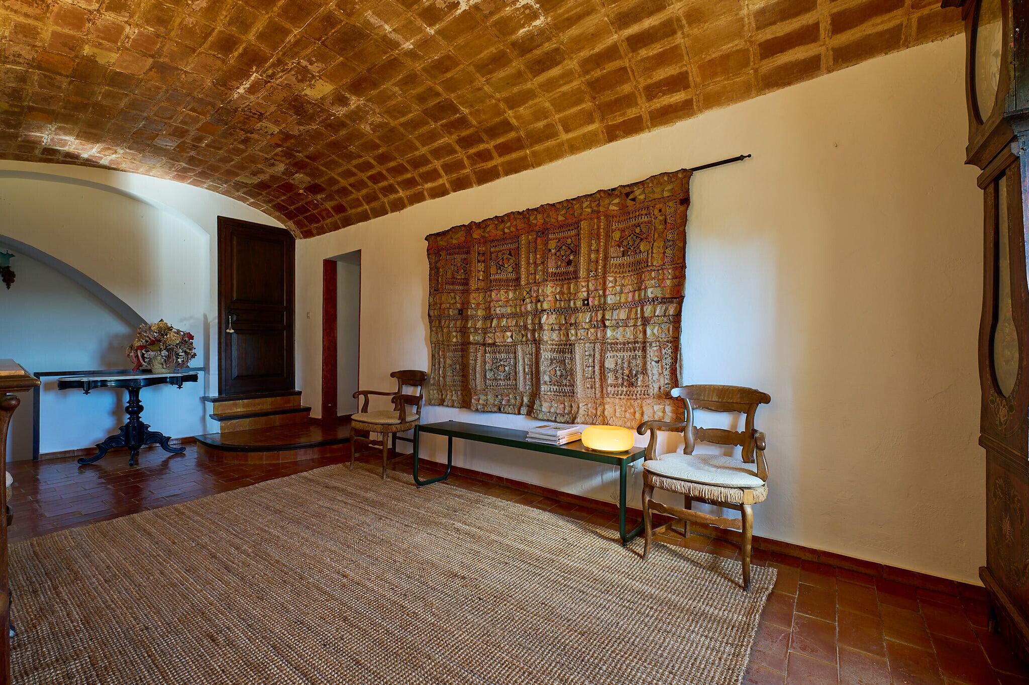 A room with a high ceiling, a patterned tapestry, a wooden bench, two chairs, and a small table.