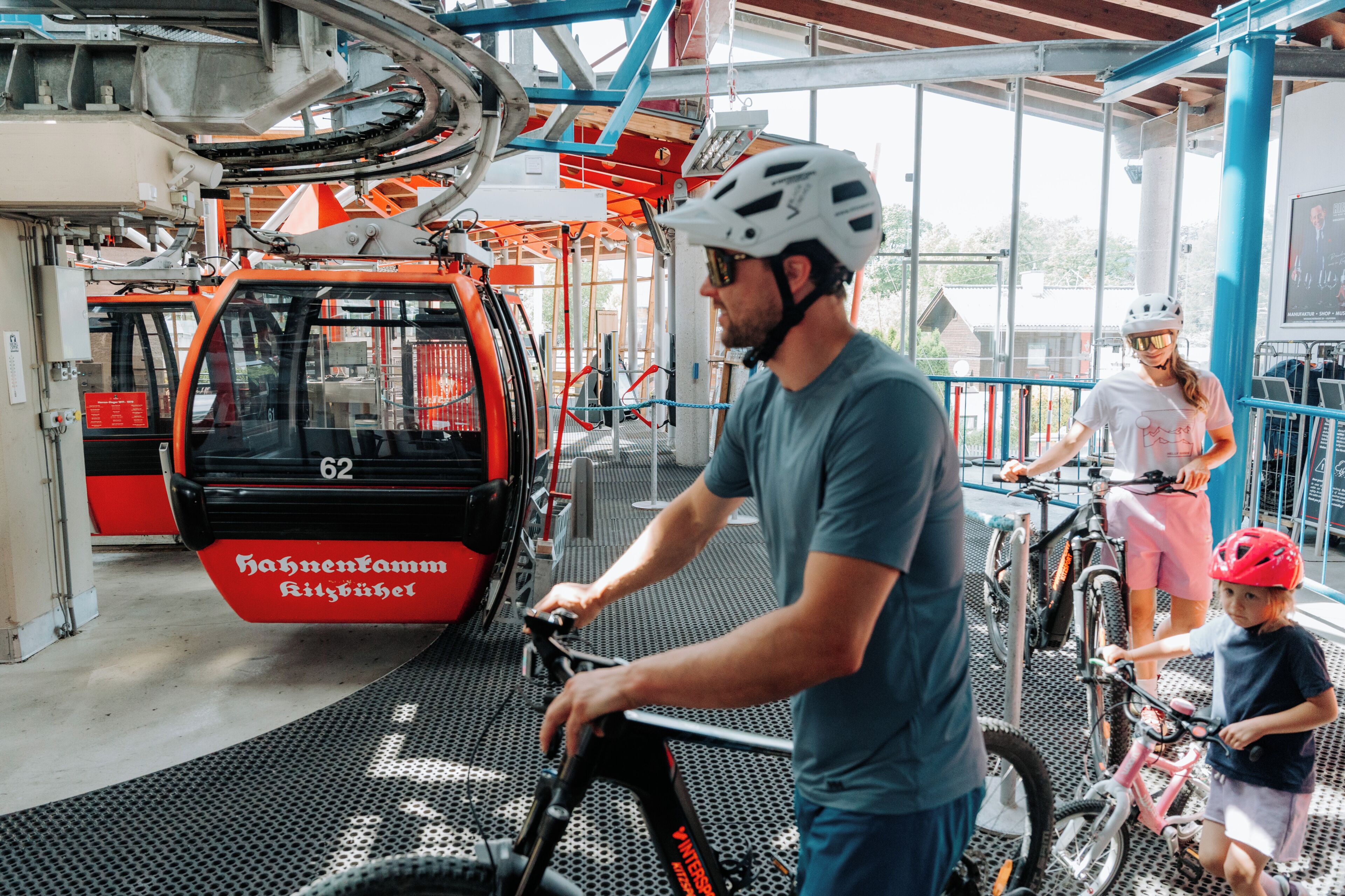 Un hombre en bicicleta, una mujer en bicicleta y un niño en bicicleta en un espacio cubierto, con un teleférico al fondo.