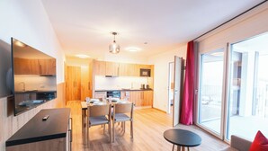 A modern kitchen with wooden cabinets, a dining area with a table and chairs, and a balcony with a red curtain. - KitzSki Living (Kitzbühel)