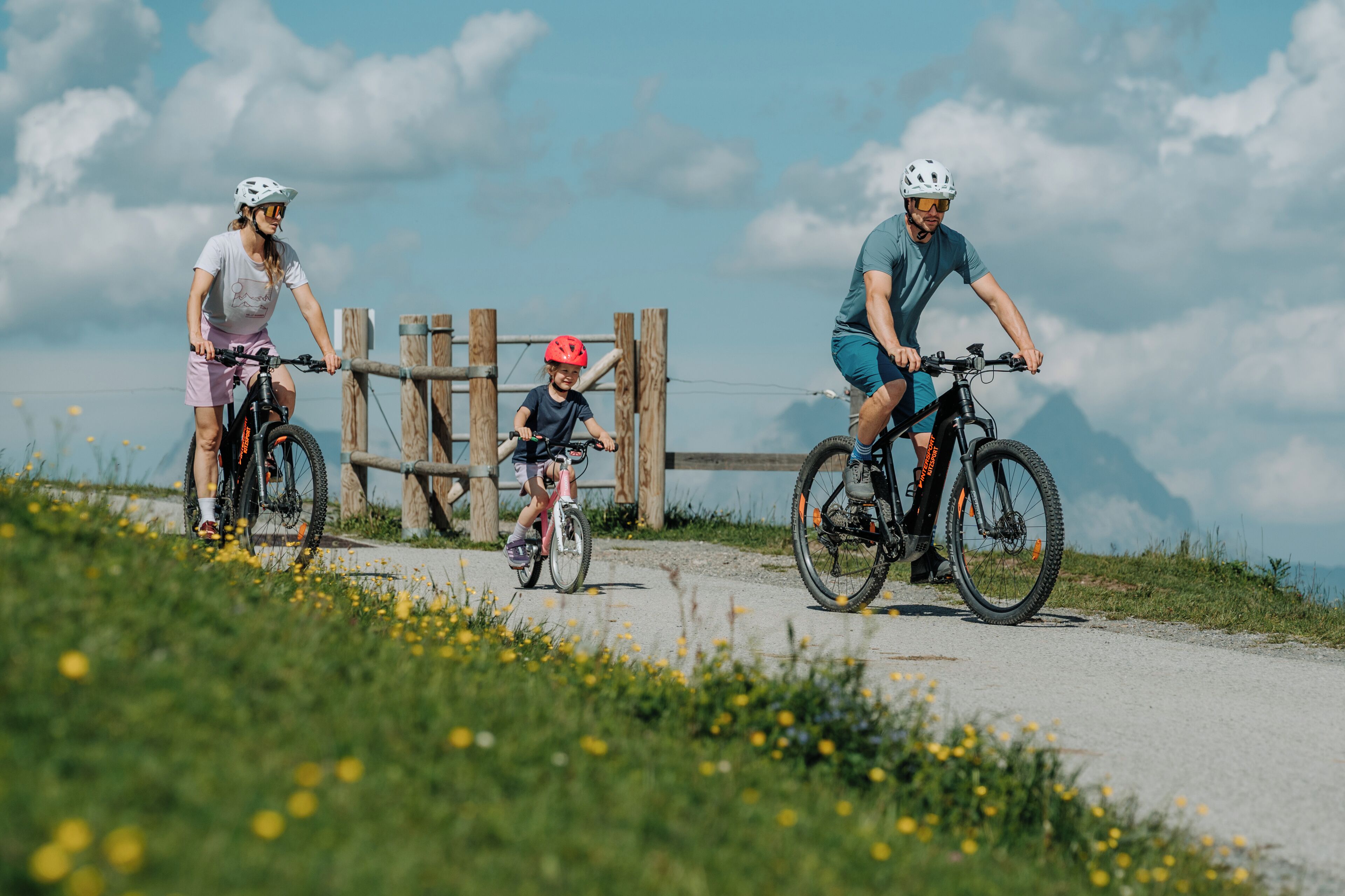 Una familia en un sendero para bicicletas de montaña, con una cerca de madera y montañas al fondo.
