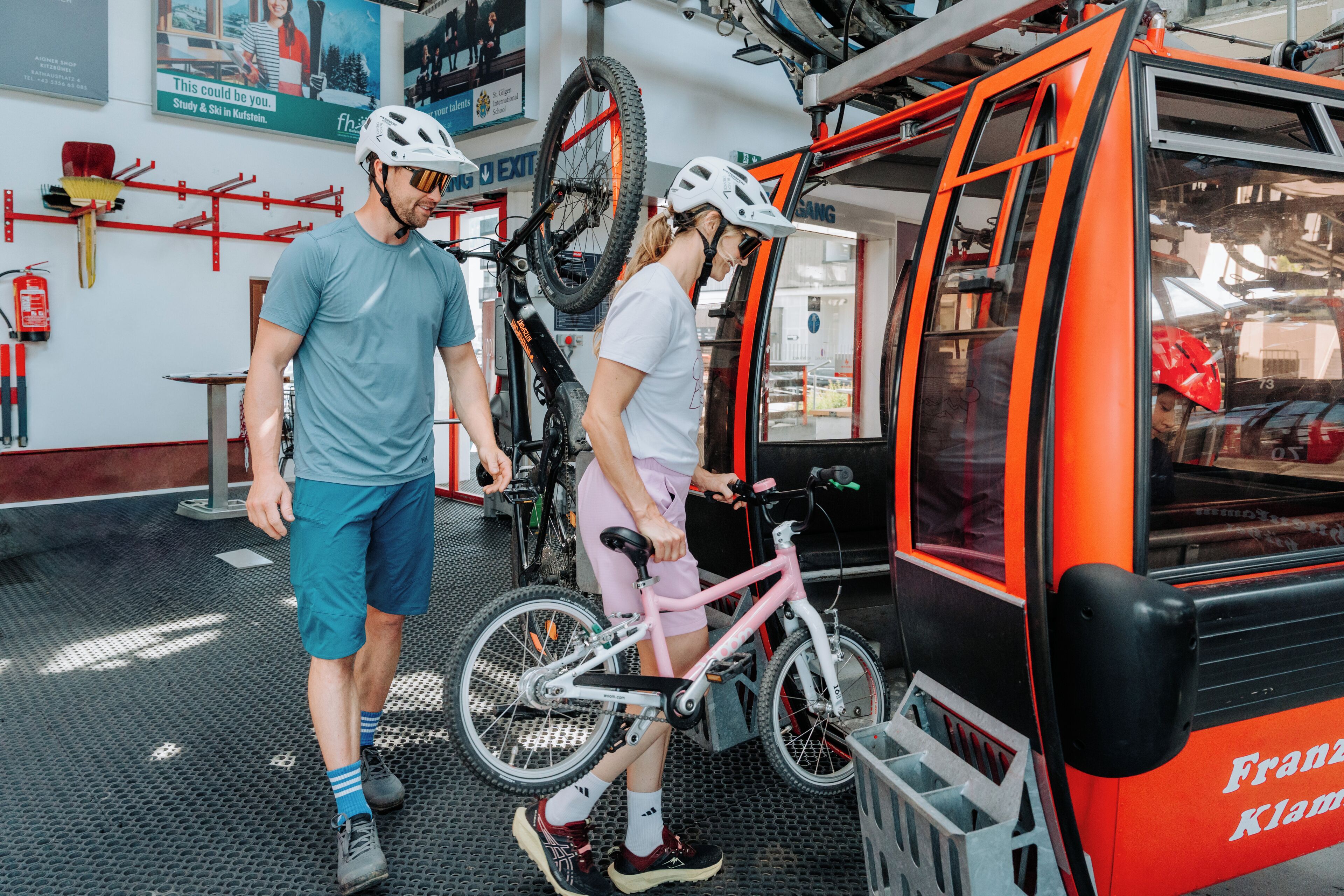 A man and a woman standing near a bicycle and a red vehicle, possibly a tram or bus, in an indoor setting with a tiled floor and various equipment and tools on the walls.