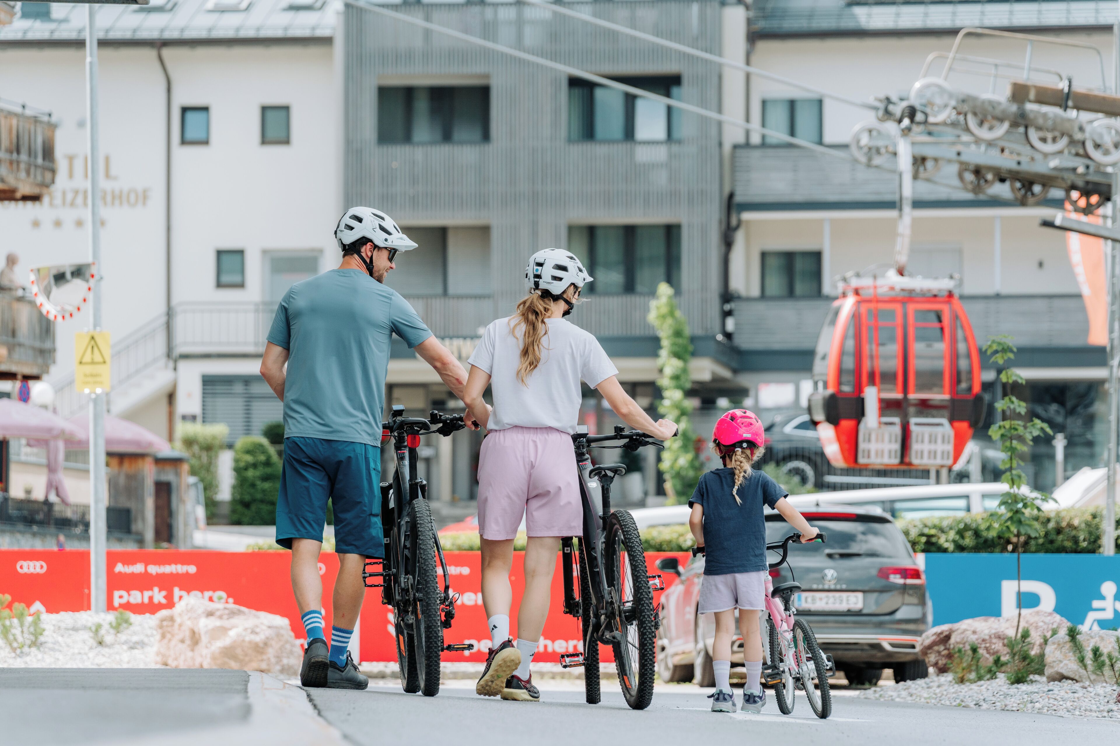 Een groep mensen met fietsen, een rood voertuig en een gebouw met een bord waarop staat 'AUDI QUATTRO parkeer hier'.