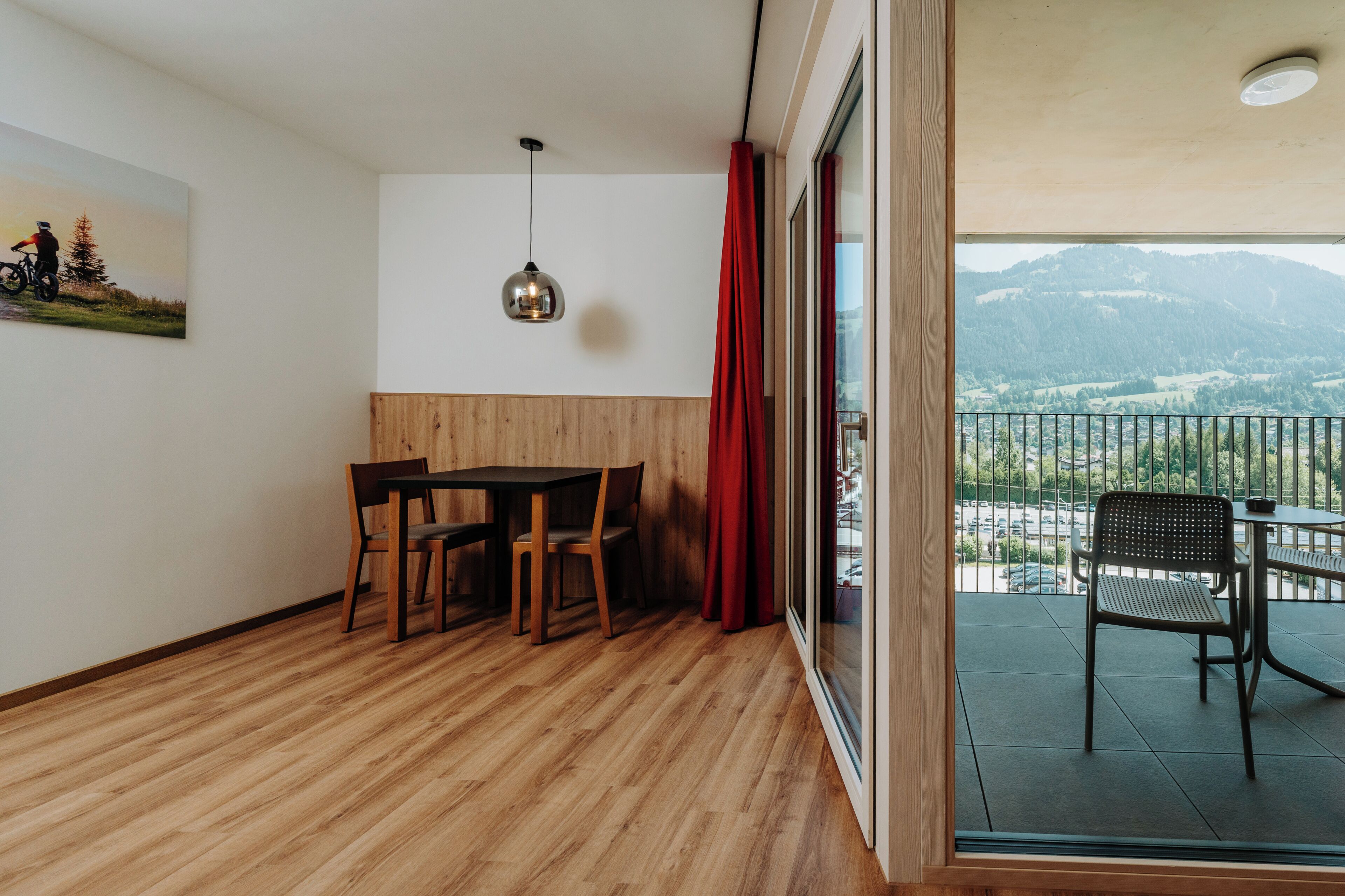 Una habitación con suelo de madera, una mesa de comedor con sillas, una lámpara colgante, una cortina roja y un balcón con vistas a montañas.