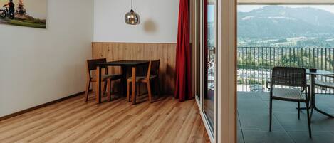A room with a wooden floor, a dining table with chairs, a pendant light, a red curtain, and a balcony with a view of mountains.