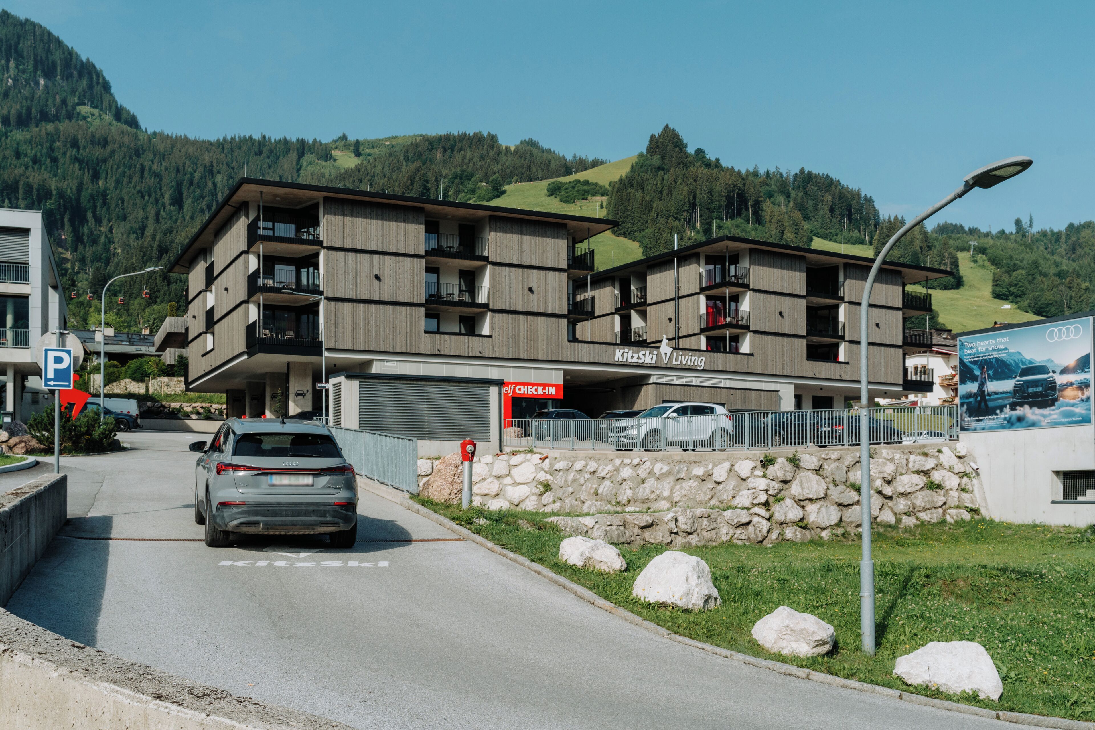 A multi-story building with balconies, a parking area, and a mountain backdrop.