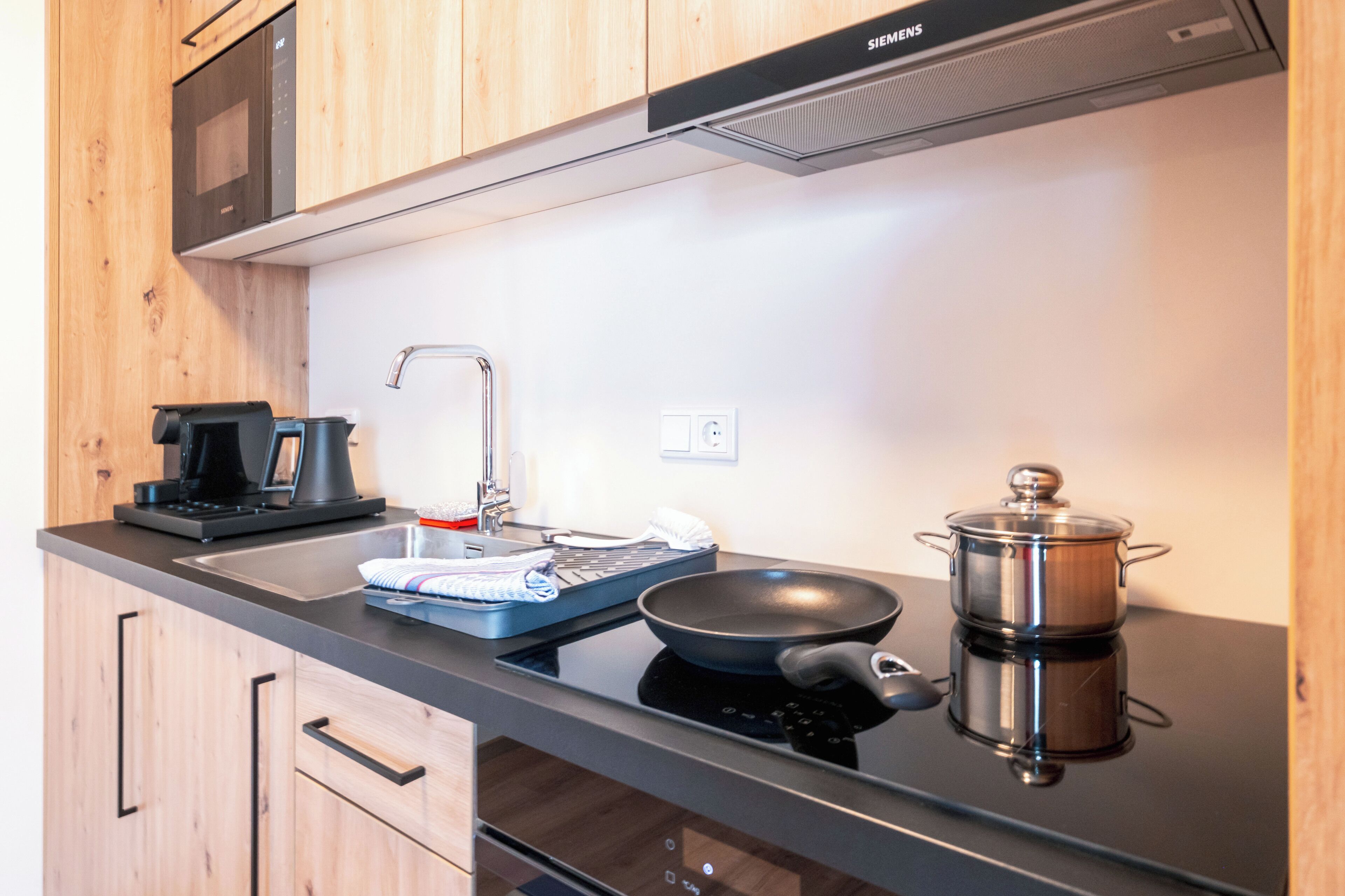 Een moderne keuken met houten kastjes, een zwart aanrecht, een magnetron, een koffiezetapparaat, een spoelbak en een kookplaat met een pan.