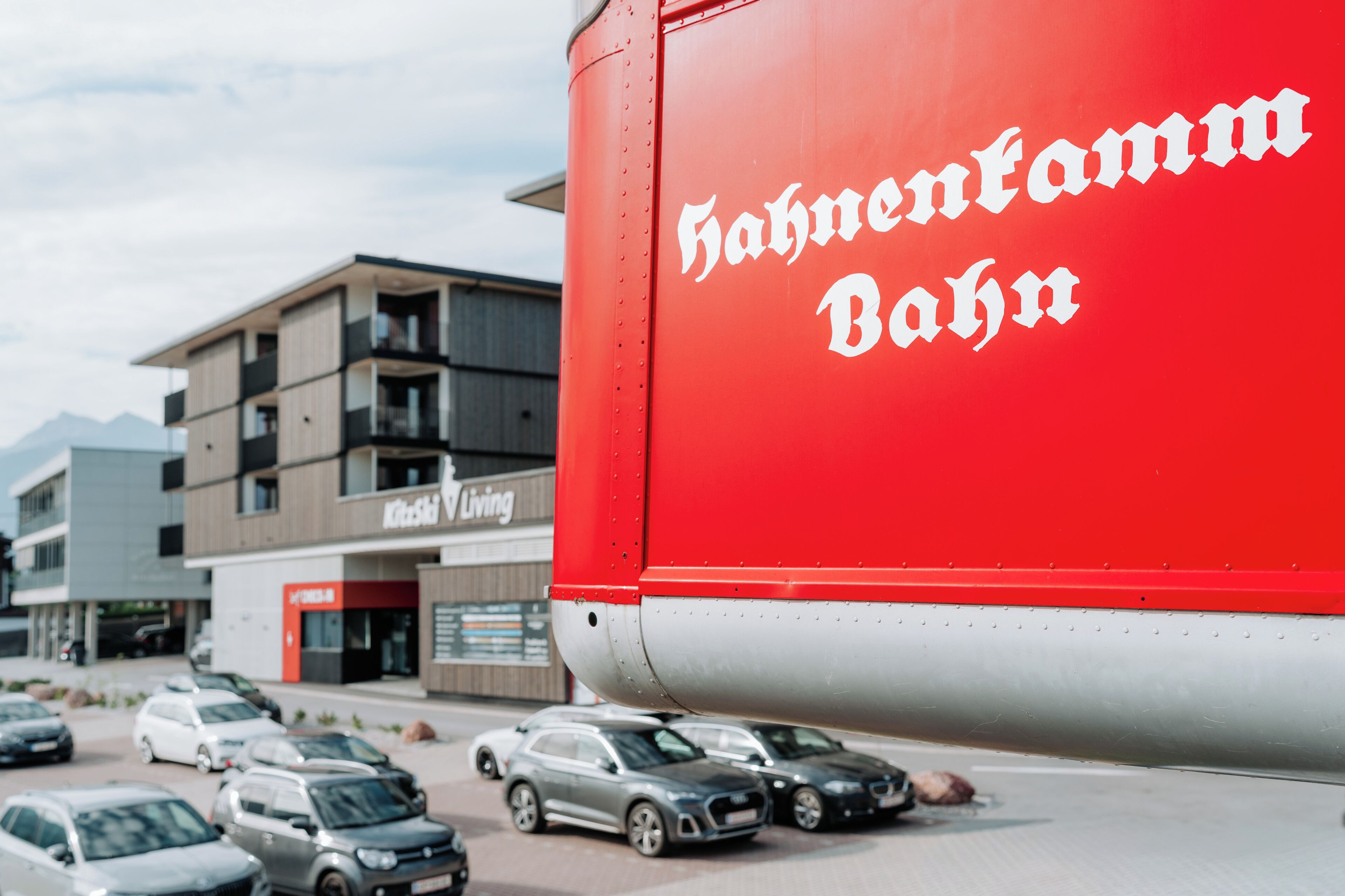 A red truck with 'Hahnentamm Bahn' written on it parked in a parking lot with cars and a building in the background.