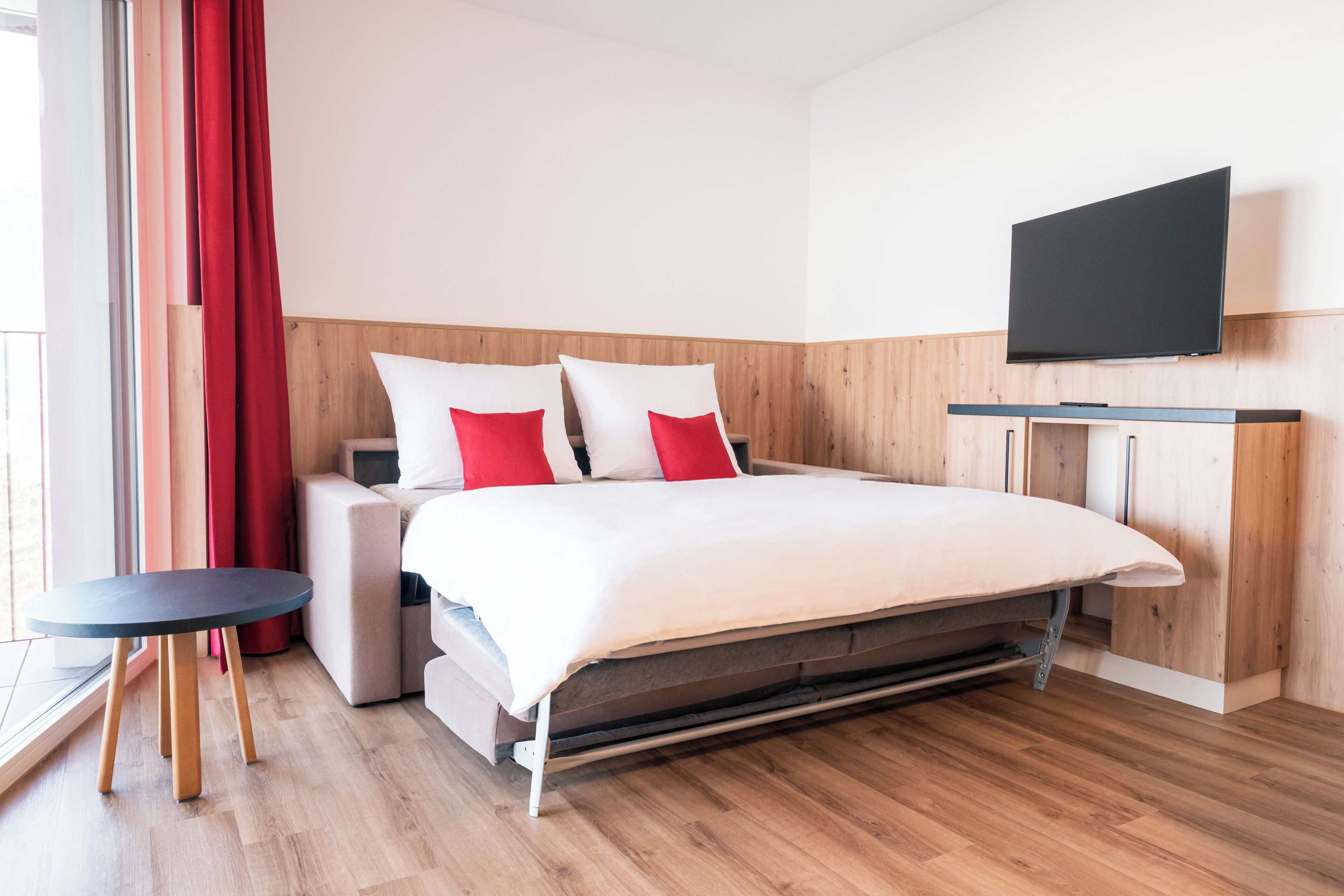 A modern hotel room with a bed, a TV, a small table, and a window with curtains.