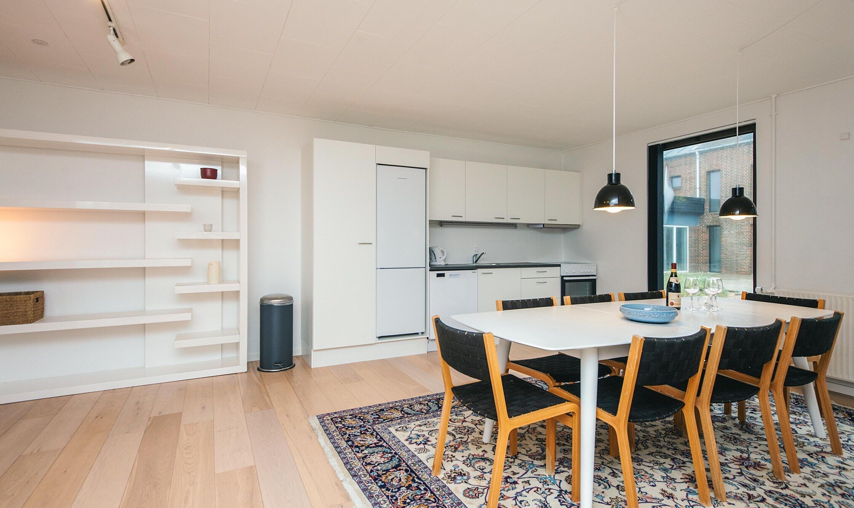Une cuisine moderne avec des meubles blancs, un coin repas comprenant une table blanche et des chaises noires, et un tapis à motifs sur le sol.