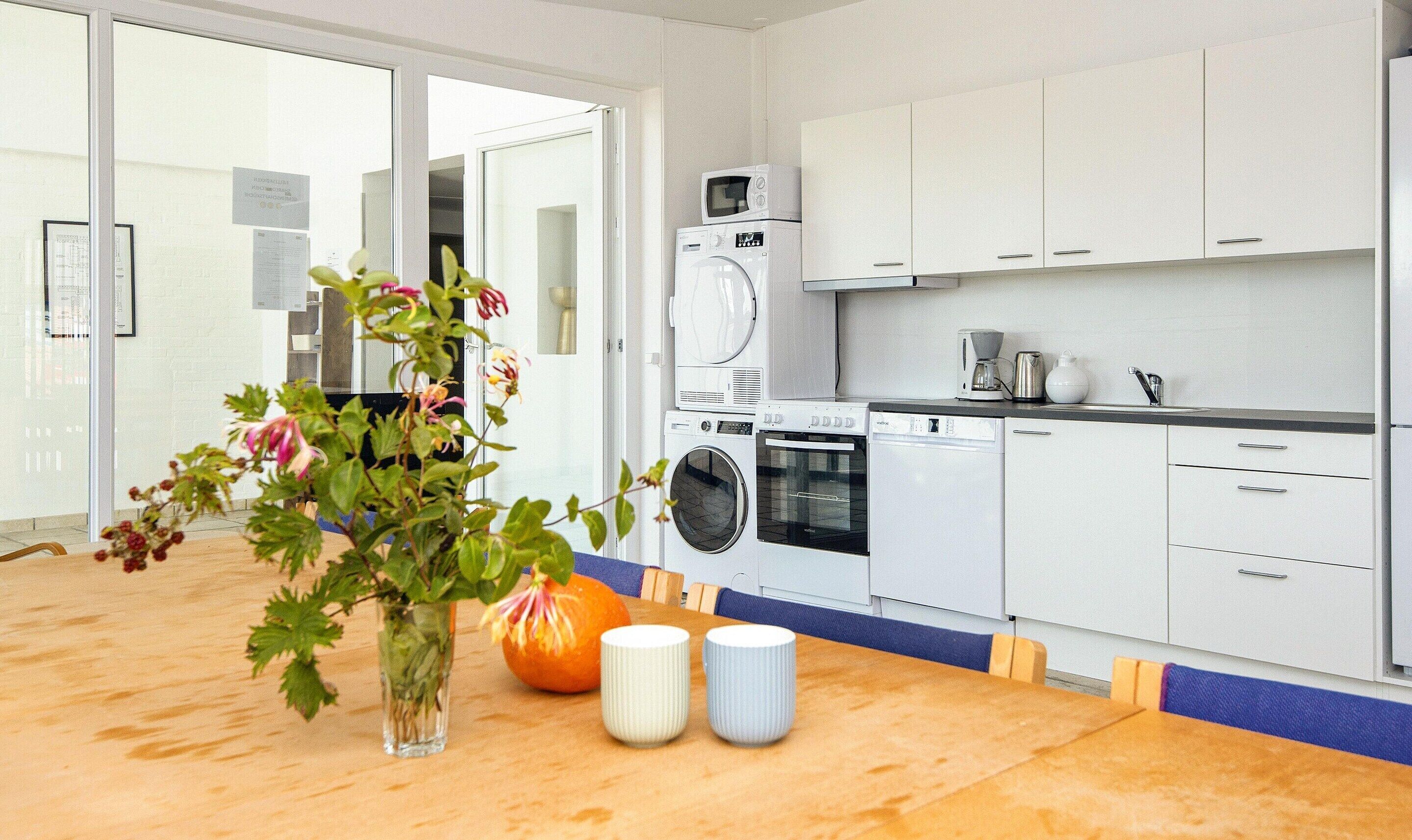 A modern kitchen with white cabinets, a built-in oven, microwave, and dishwasher. There is a wooden table with a vase of flowers and two mugs.