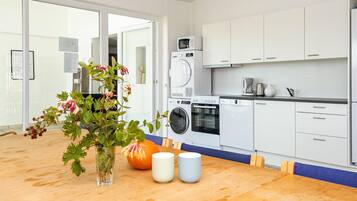 A modern kitchen with white cabinets, a built-in oven, microwave, and dishwasher. There is a wooden table with a vase of flowers and two mugs.