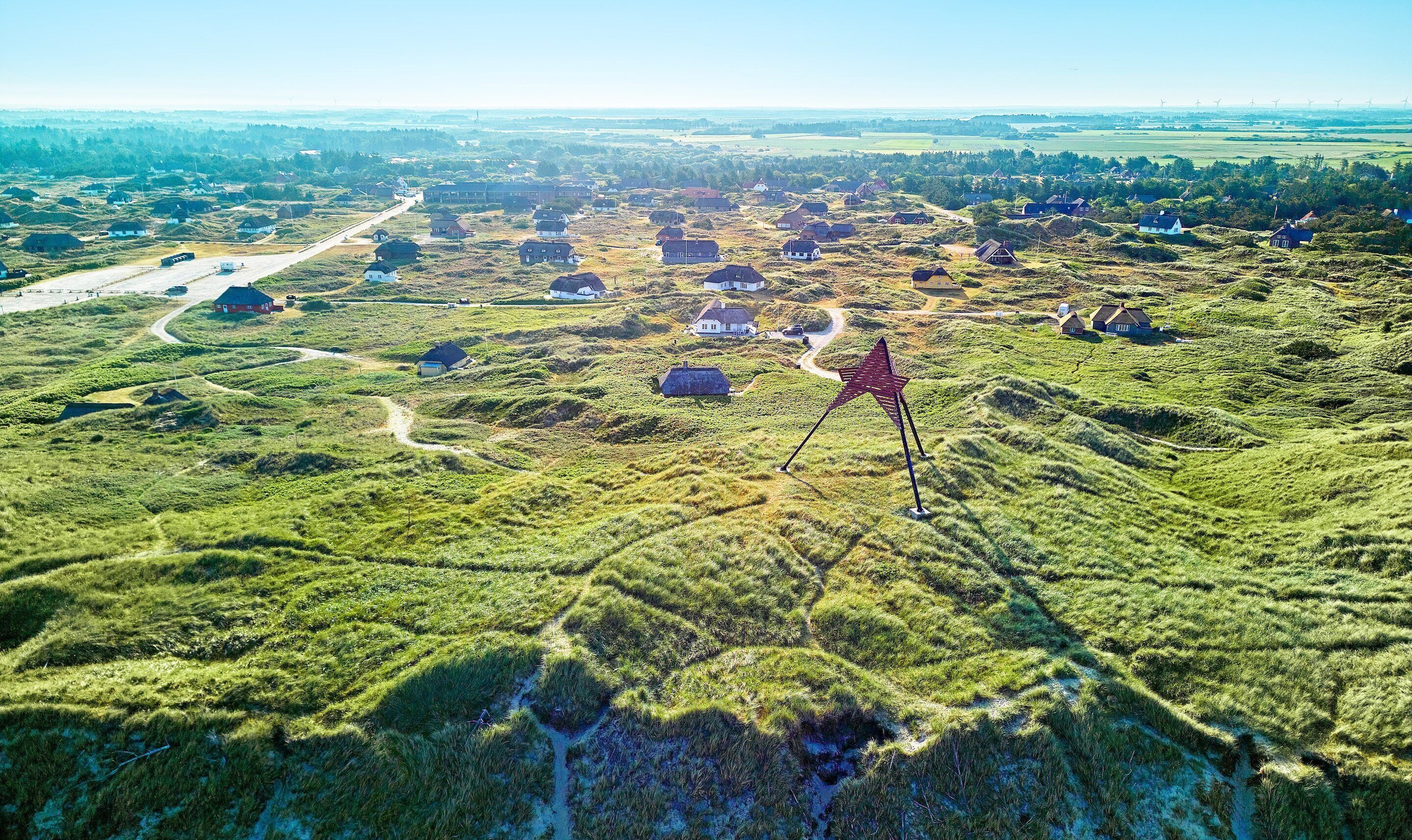 Vista aérea de un paisaje rural con casas dispersas, una estructura prominente en forma de estrella roja y una carretera.