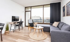Una sala de estar moderna con un sofá, dos sillones, una mesita, un televisor y un amplio ventanal con vistas a edificios.