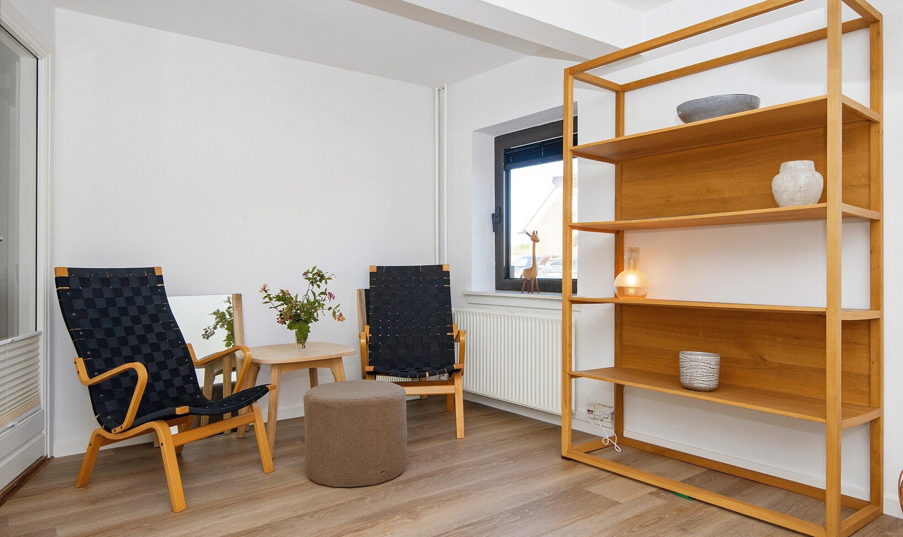Una habitación con dos sillas, una mesita pequeña, un otoman redondo y una estantería de madera.