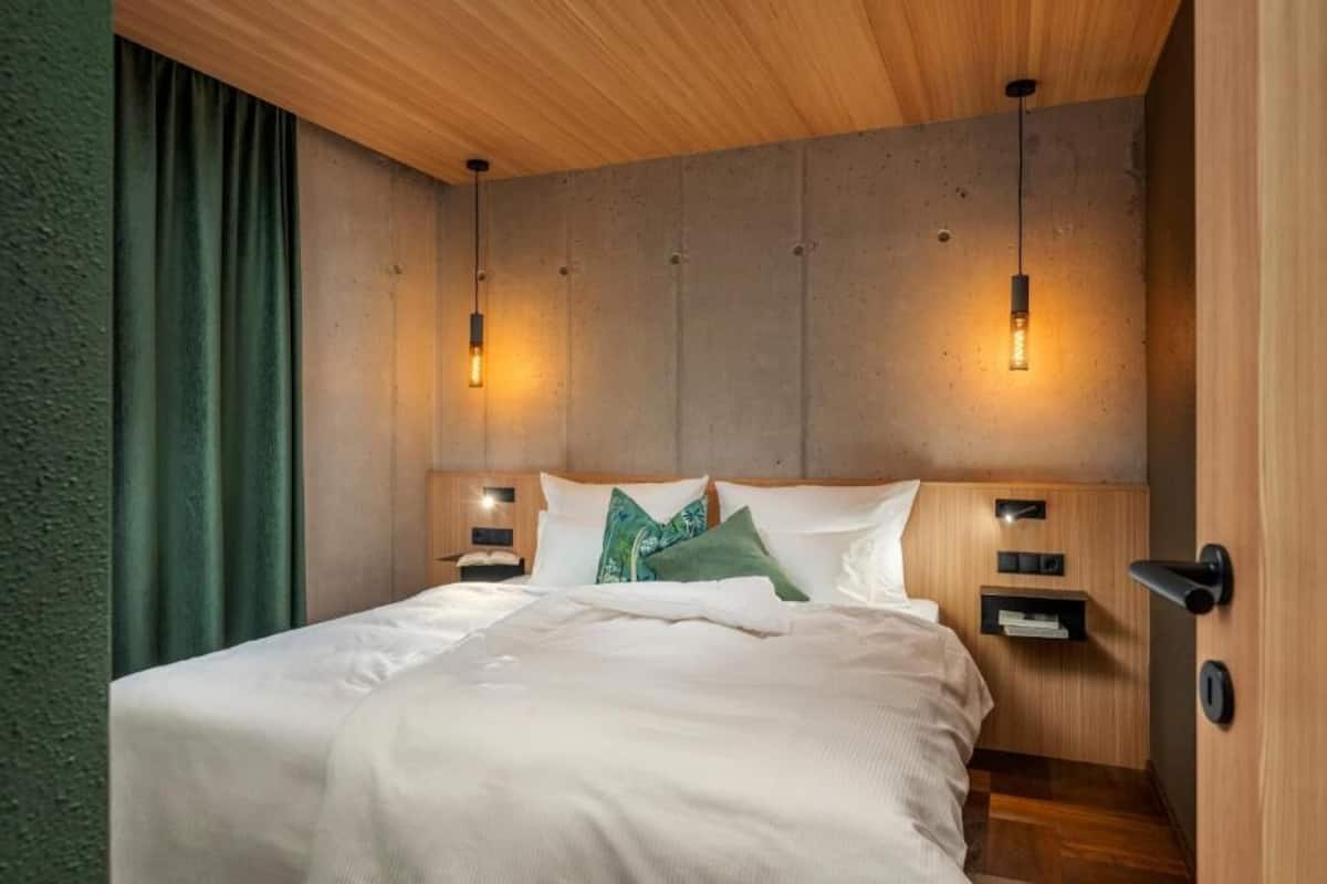 Quarto de hotel moderno com teto de madeira, uma cama grande com roupa de cama branca, duas mesas de cabeceira com abajures e uma cortina verde.