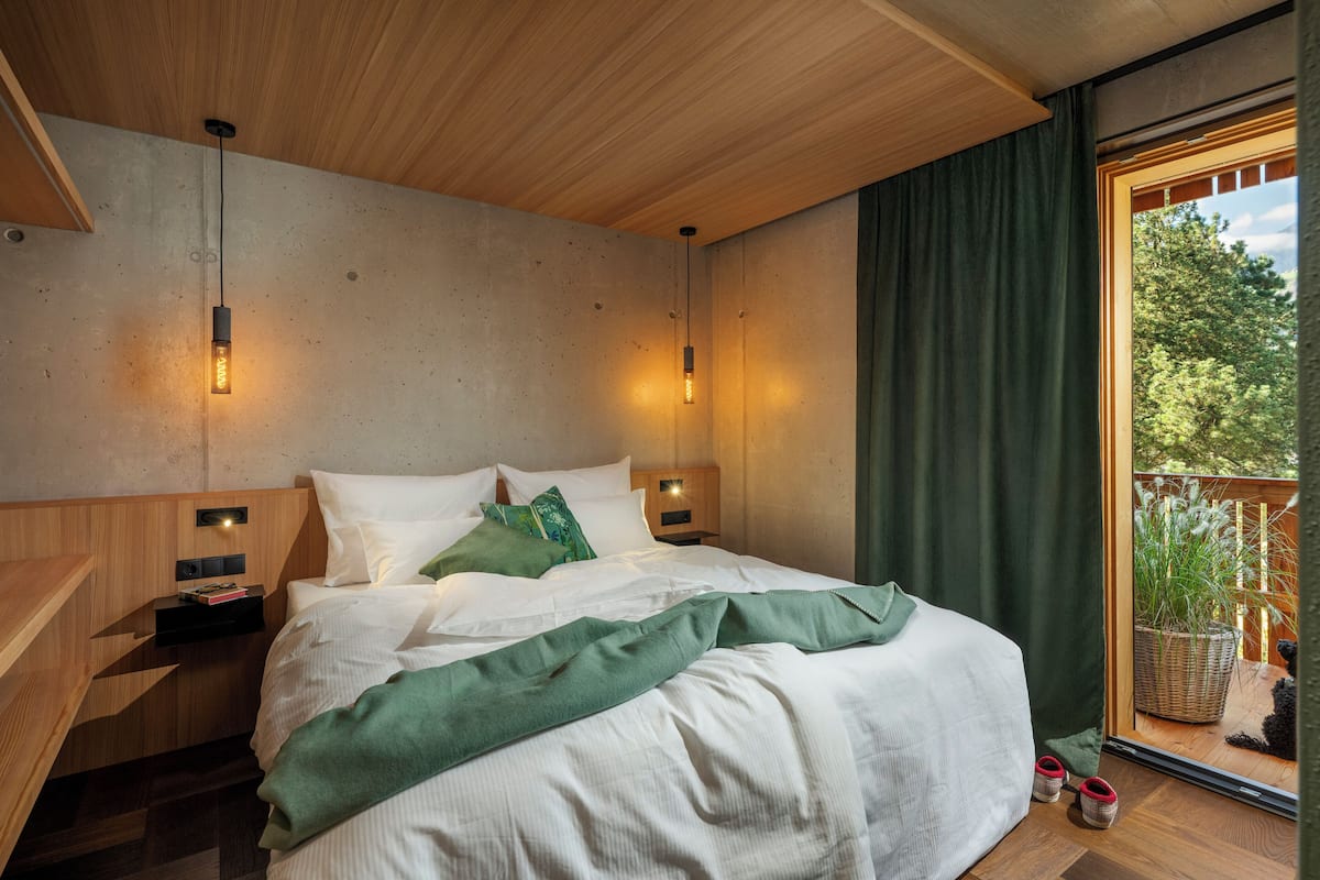 Quarto com teto de madeira, cama com roupa de cama branca, almofadas verdes e mesa de cabeceira com abajur.