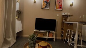 A cozy living room with a flat-screen TV, two wall-mounted light fixtures, a small wooden table with a potted plant and candle, and a dining area with a white table and chairs. - CosyWellington-Studio near Arras station and center (Achicourt)