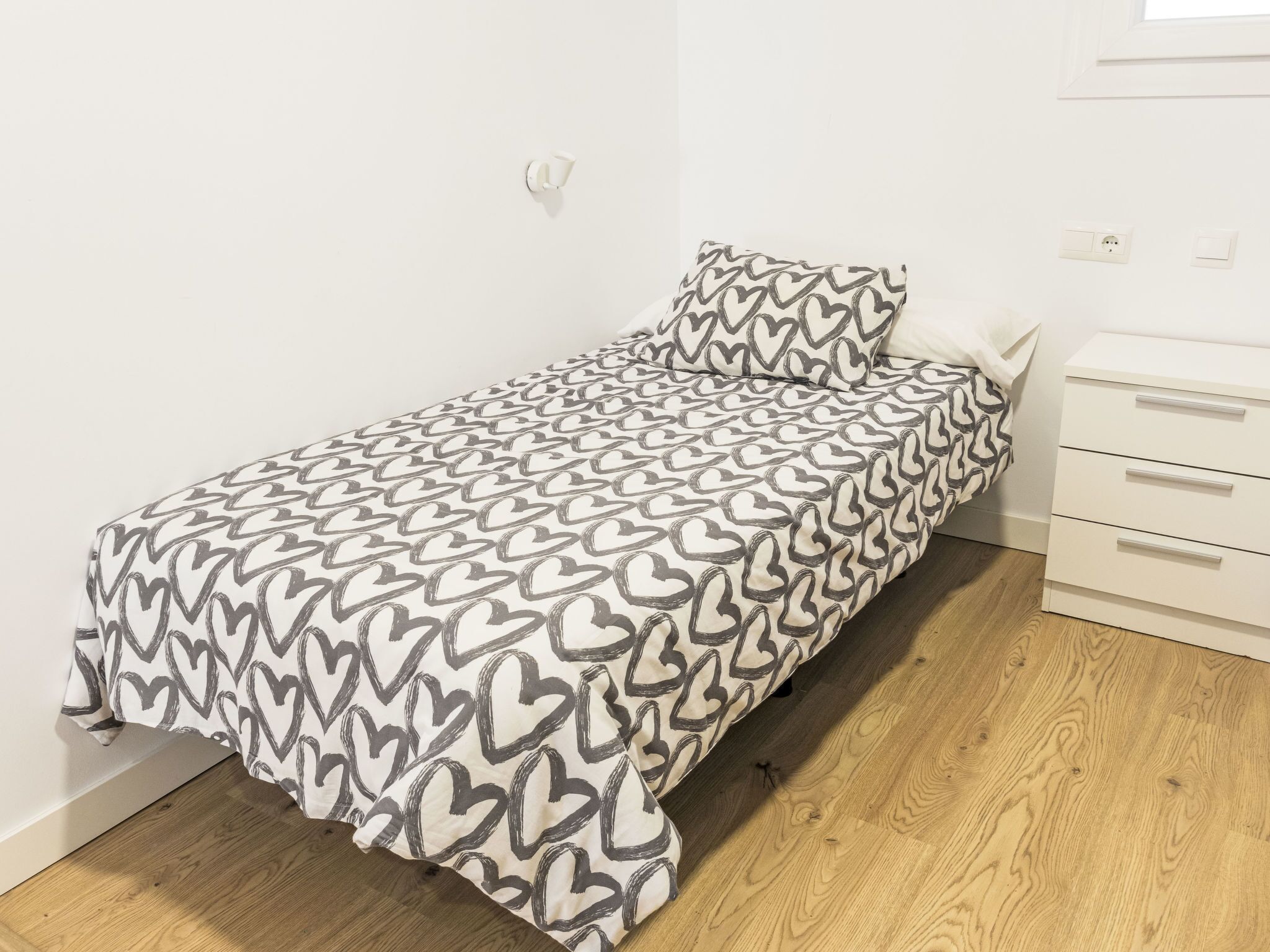 Un petit lit bien fait, avec une couvre-lit à motifs et un coussin assorti, dans une pièce simple dotée d’un plancher en bois et de murs blancs.