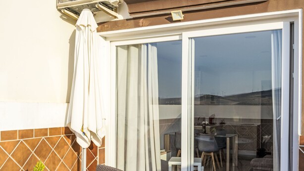 Una terrazza con tavolo in vetro, sedie in vimini, una pianta in vaso e un ombrellone bianco.