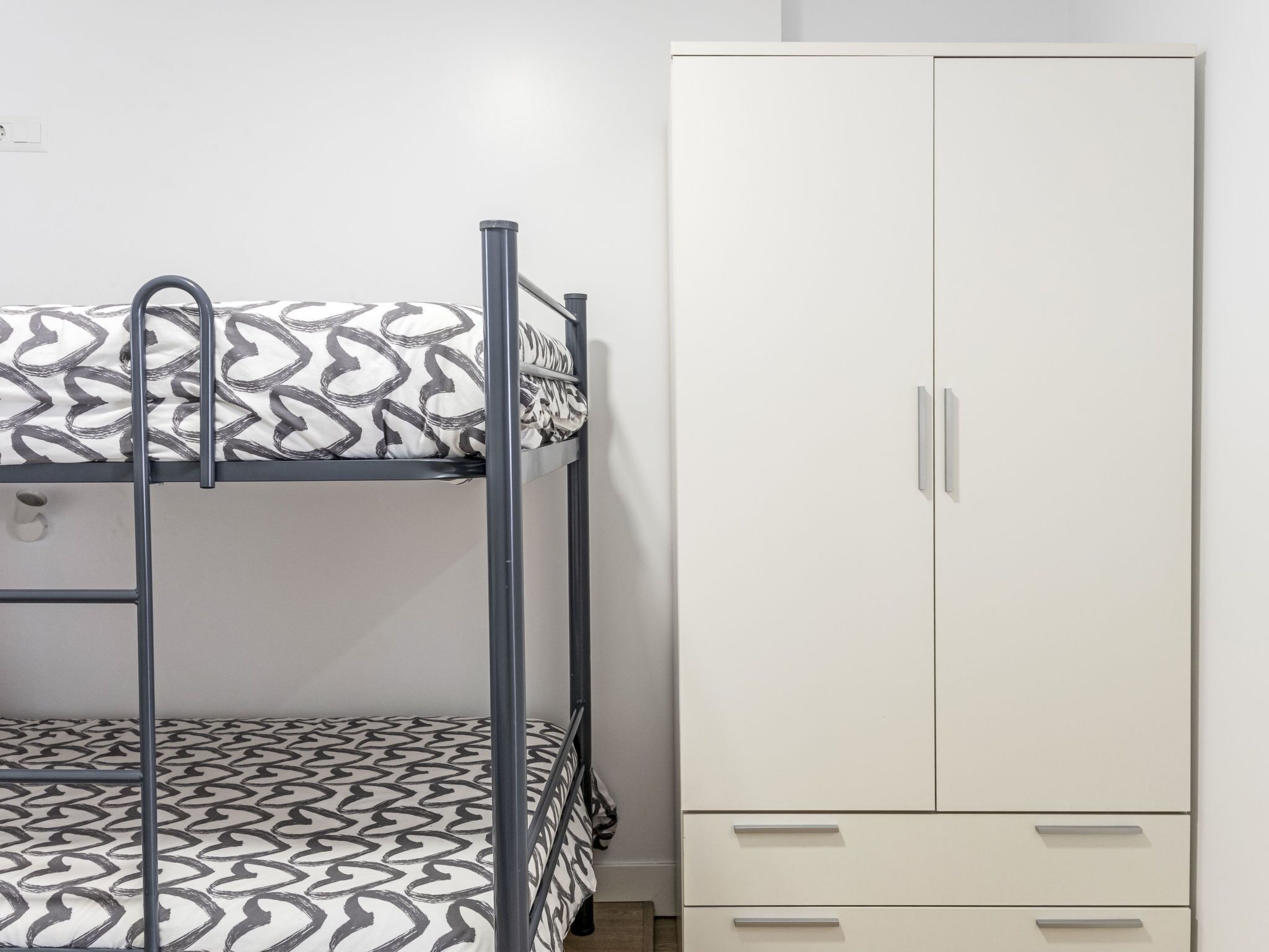 Un lit superposé avec une literie à motifs, une armoire blanche avec de nombreux tiroirs et une prise de courant au mur.