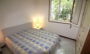 A small, simple bedroom with a single bed, a bedside lamp, and a window with a view of greenery. - Villa With Patio Near the Beach (Lignano Sabbiadoro)