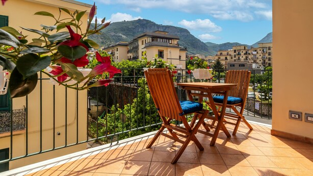 Un balcone con tavolo e sedie, affaccia su un paesaggio montuoso e una cittadina.