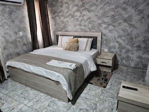 A neatly made bed with a white comforter and a folded towel on it. There is a wooden nightstand with a drawer and a small table beside the bed. The room has a patterned wallpaper and marble flooring.