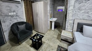 Una habitación de hotel con cama, una silla, una mesita, un armario, un lavabo y un aire acondicionado.