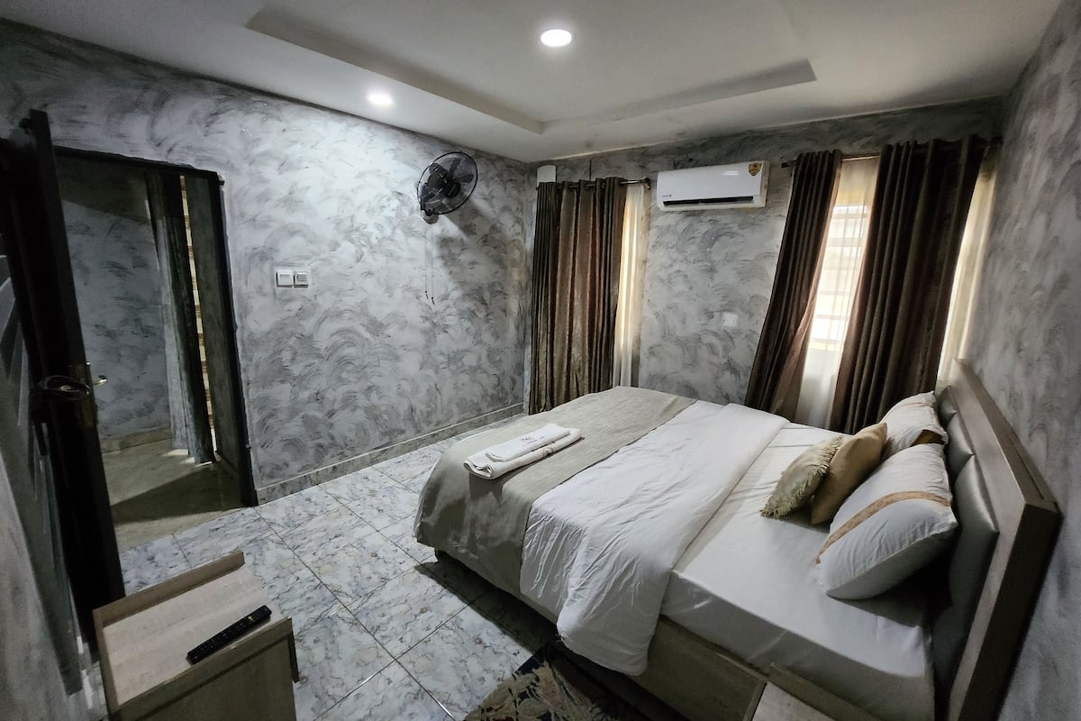 A hotel room with a bed, a bedside table, a mirror, an air conditioning unit, and a door.
