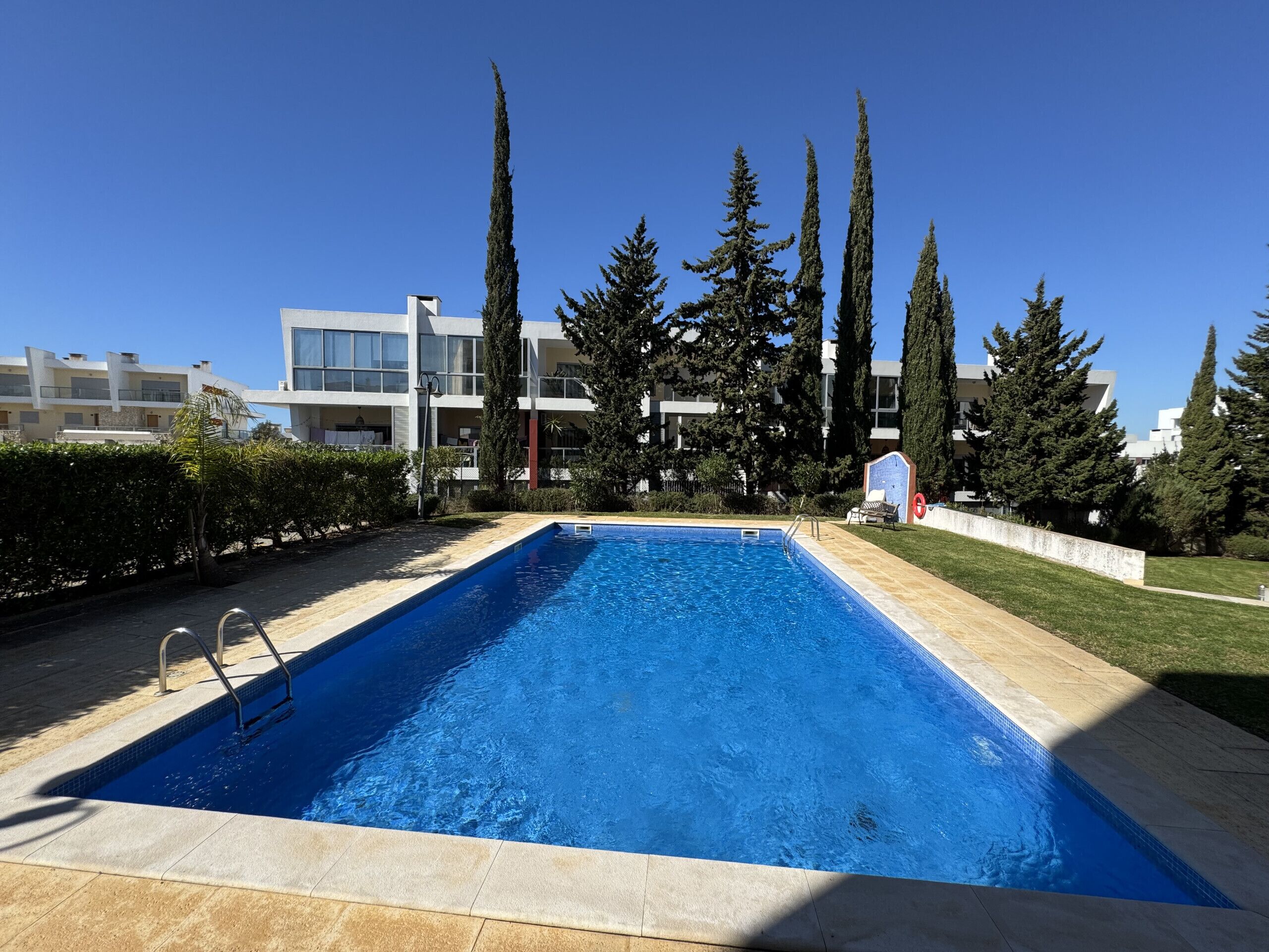 A rectangular swimming pool with a tiled edge, surrounded by a paved deck. In the background, there are modern buildings with balconies and a row of tall, slender trees.