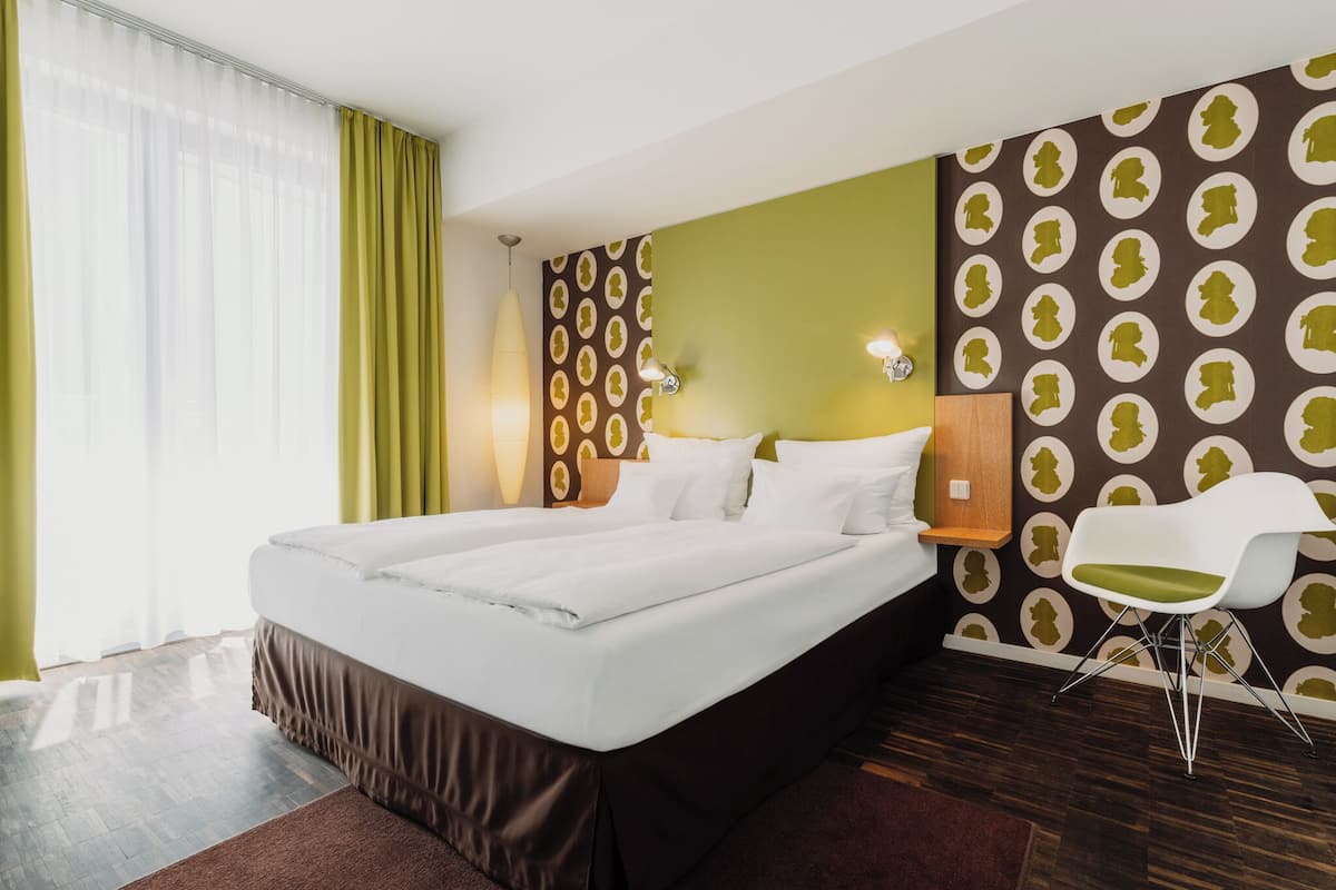 A hotel room with a large bed, a chair, a wall with a patterned design, and a window with curtains.