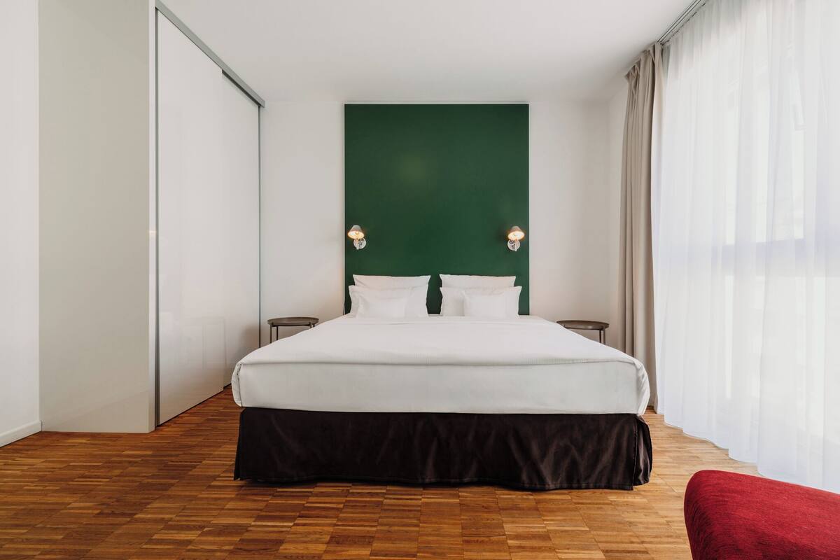 A hotel room with a large bed, a green accent wall, two bedside tables, and a red chair.