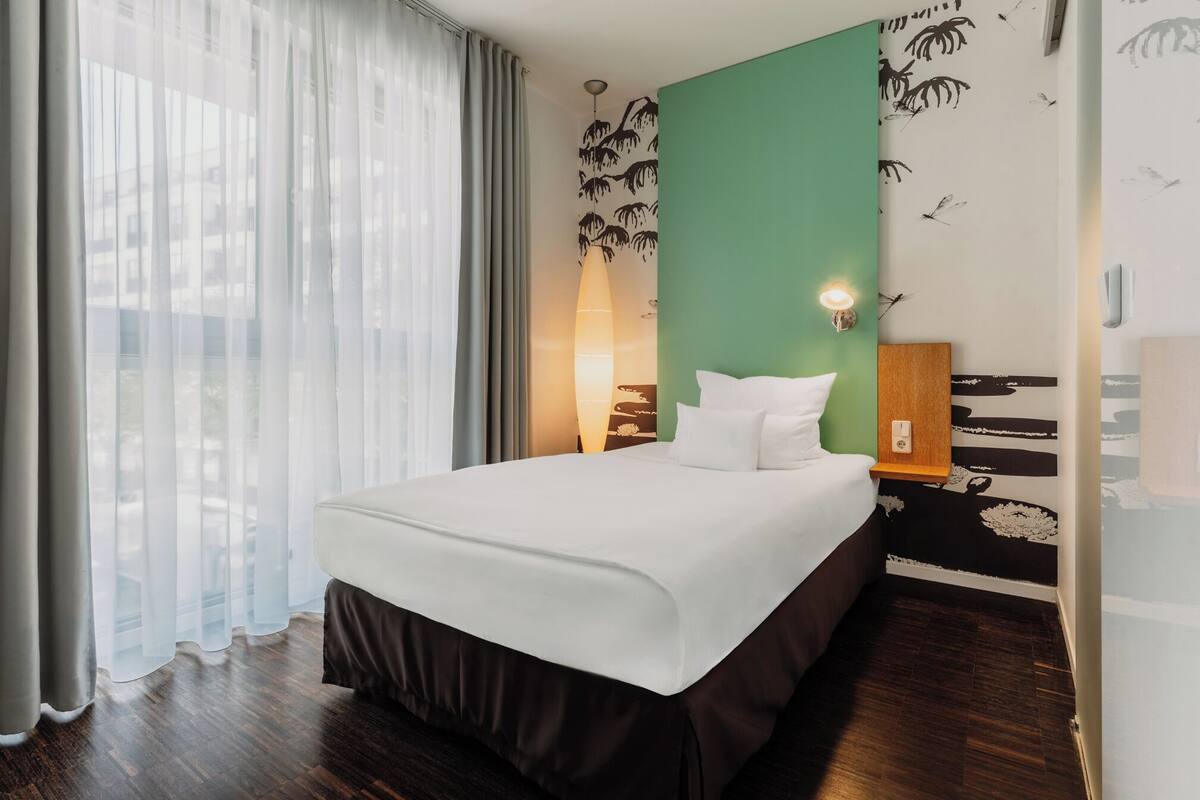 A hotel room with a bed, a nightstand, a wall-mounted lamp, and a view through sheer curtains.