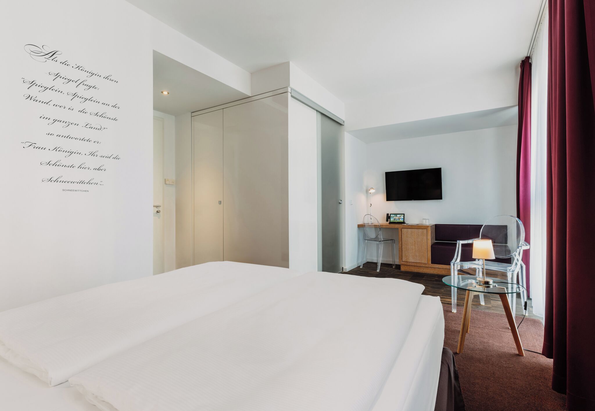 Habitació d'hotel amb un llit, una taula de treball amb una cadira, un televisor penjat a la paret i una partició de vidre.