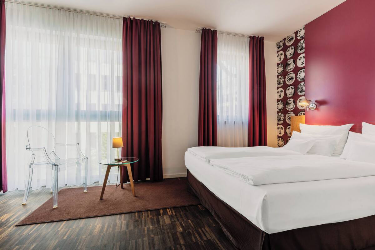 A hotel room with a bed, a small table with a lamp, a chair, and a view of the city through sheer curtains.