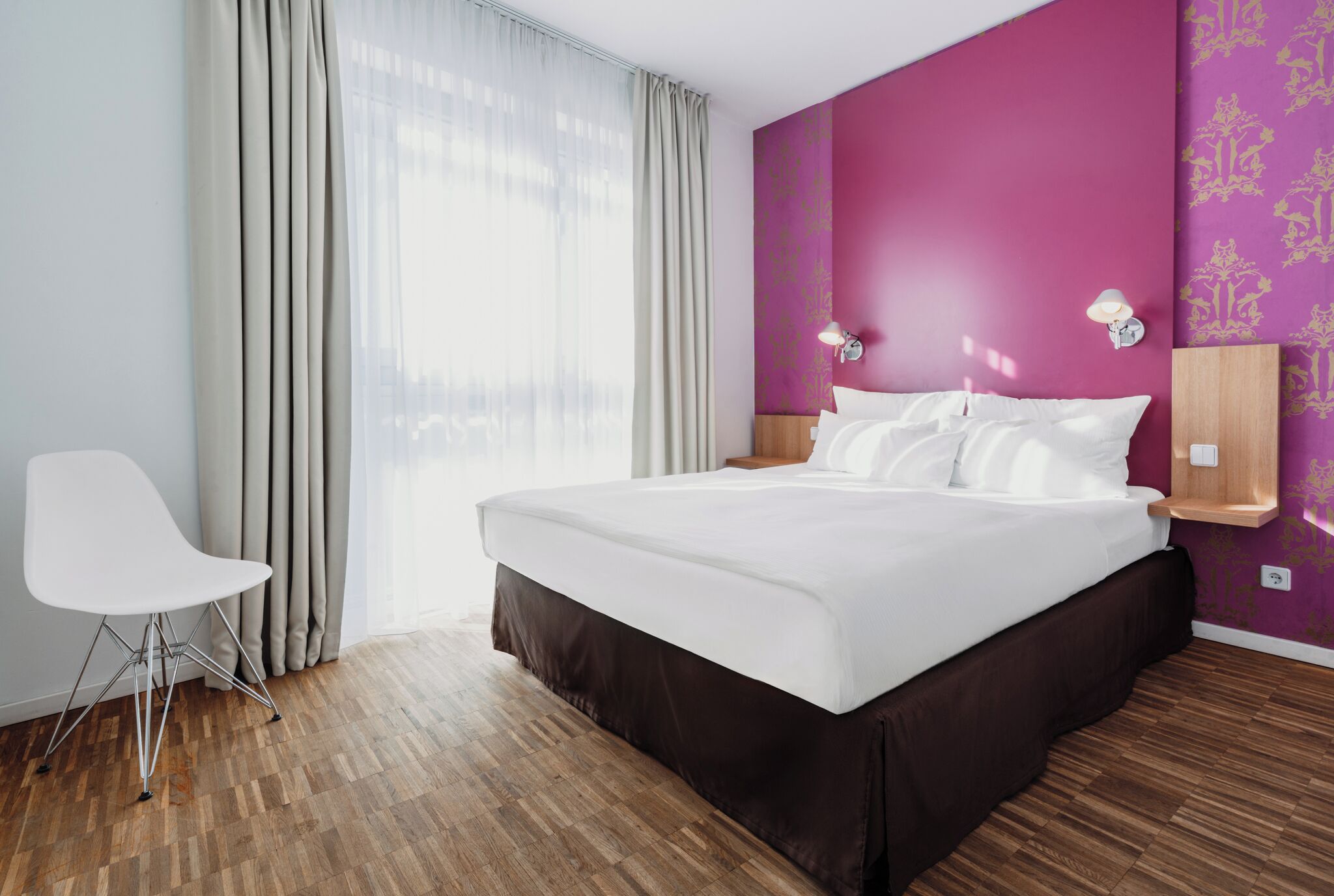A hotel room with a large bed, a chair, a window with curtains, and a wall with a patterned wallpaper.
