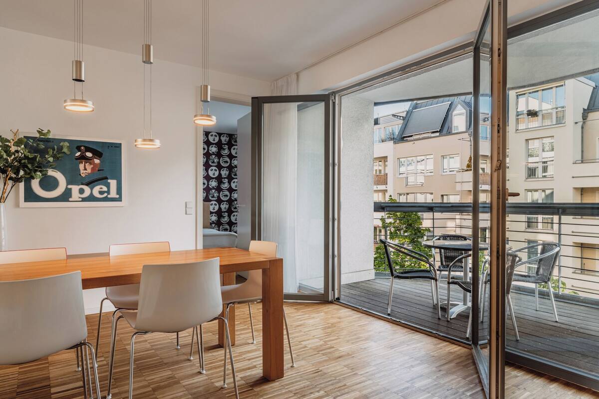 A modern dining area with a wooden table and chairs, a large window with a view of buildings, and pendant lights.