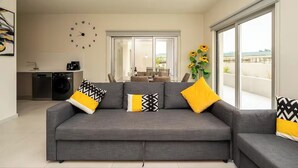 Una sala de estar moderna con un sofá gris, cojines estampados en negro y amarillo, un reloj de pared y un ventanal con vistas al exterior.
