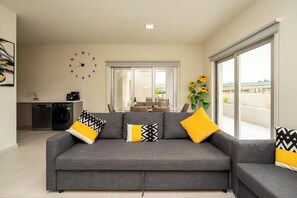 A modern living room with a grey sofa, black and yellow patterned cushions, a wall clock, and a large window with a view of the outdoors.