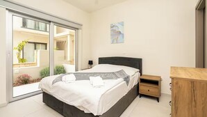 Un dormitorio con cama, una mesita de noche, un aparador de madera, un cuadro en la pared y un ventanal corredero que da a un balcón con plantas.
