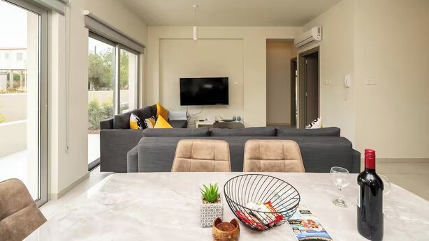 Una sala de estar moderna con un sofá grande, una mesa de comedor con una botella de vino y un televisor montado en la pared.
