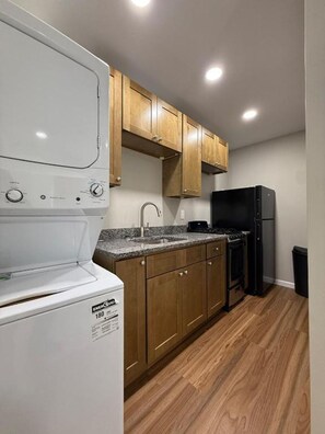 Cocina compacta con gabinetes de madera, lavadora blanca, refrigerador negro y encimera de granito.