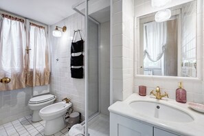 A modern bathroom with white tiles, a toilet, a bidet, a sink with a gold faucet, and a mirror with a light fixture. - Liberty Trastevere House (Rome)