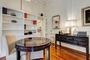 A room with a round wooden table, a white chair, a bookshelf with various items, a mirror, a lamp, and a small kitchen area with a toaster. - Liberty Trastevere House (Rome)