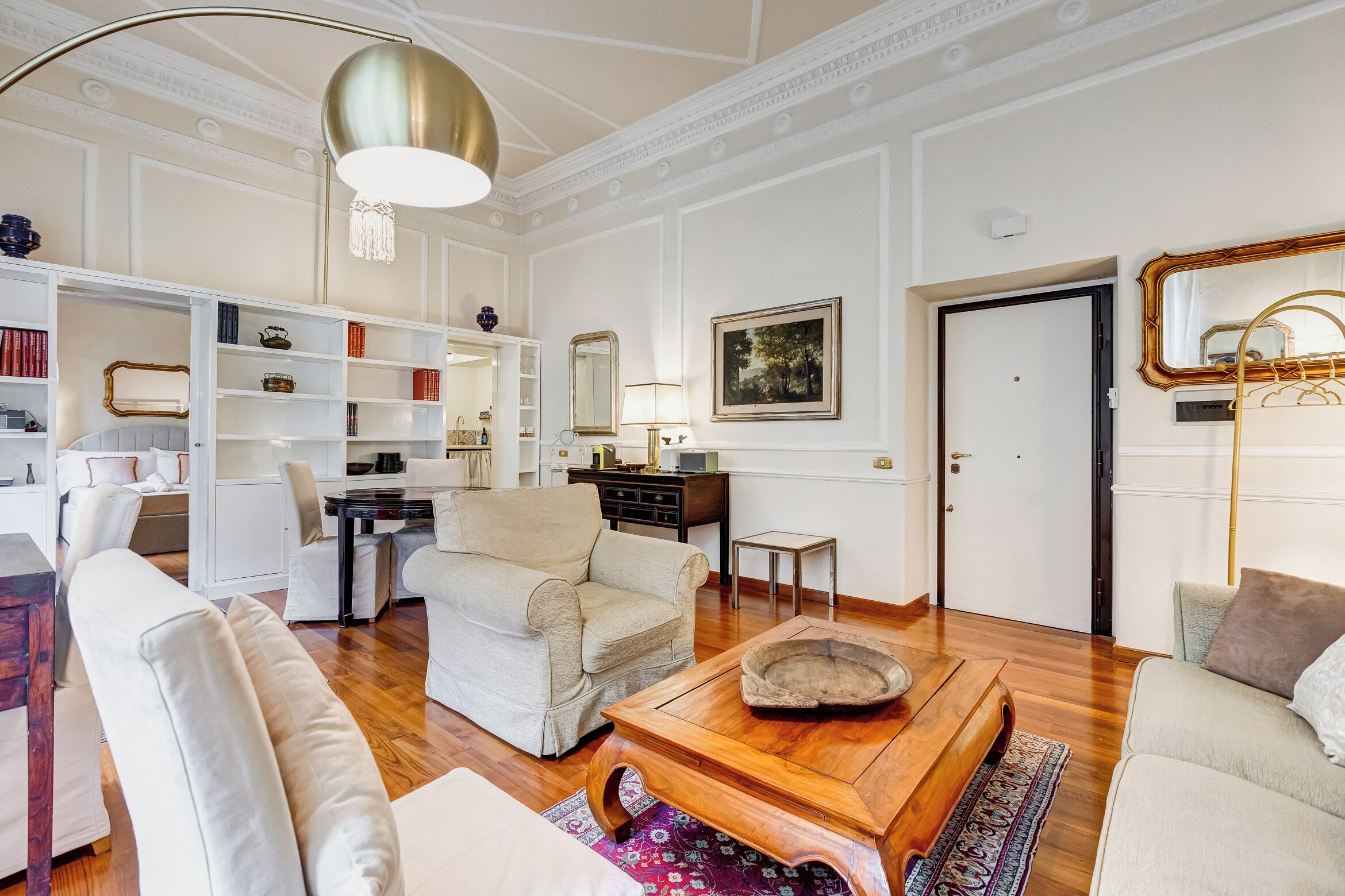 A spacious living room with a wooden coffee table, a beige sofa, and a white armchair. There is a bookshelf filled with books and decorative items, a mirror, and a door leading to another room.