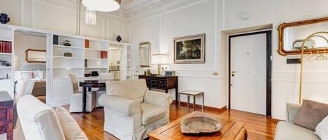 A spacious living room with a wooden coffee table, a beige sofa, and a white armchair. There is a bookshelf filled with books and decorative items, a mirror, and a door leading to another room.