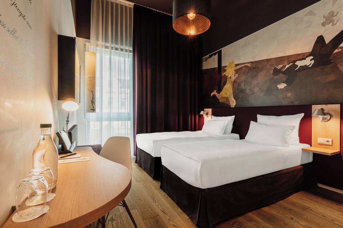 Habitación de hotel con dos camas, un escritorio con silla, una mesa con vasos y una pared decorada con un cuadro geométrico.