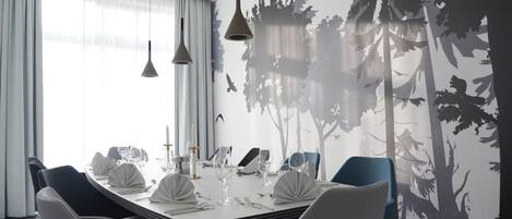 A modern dining room with a long table set for a meal, pendant lights, and a forest-themed wall mural.